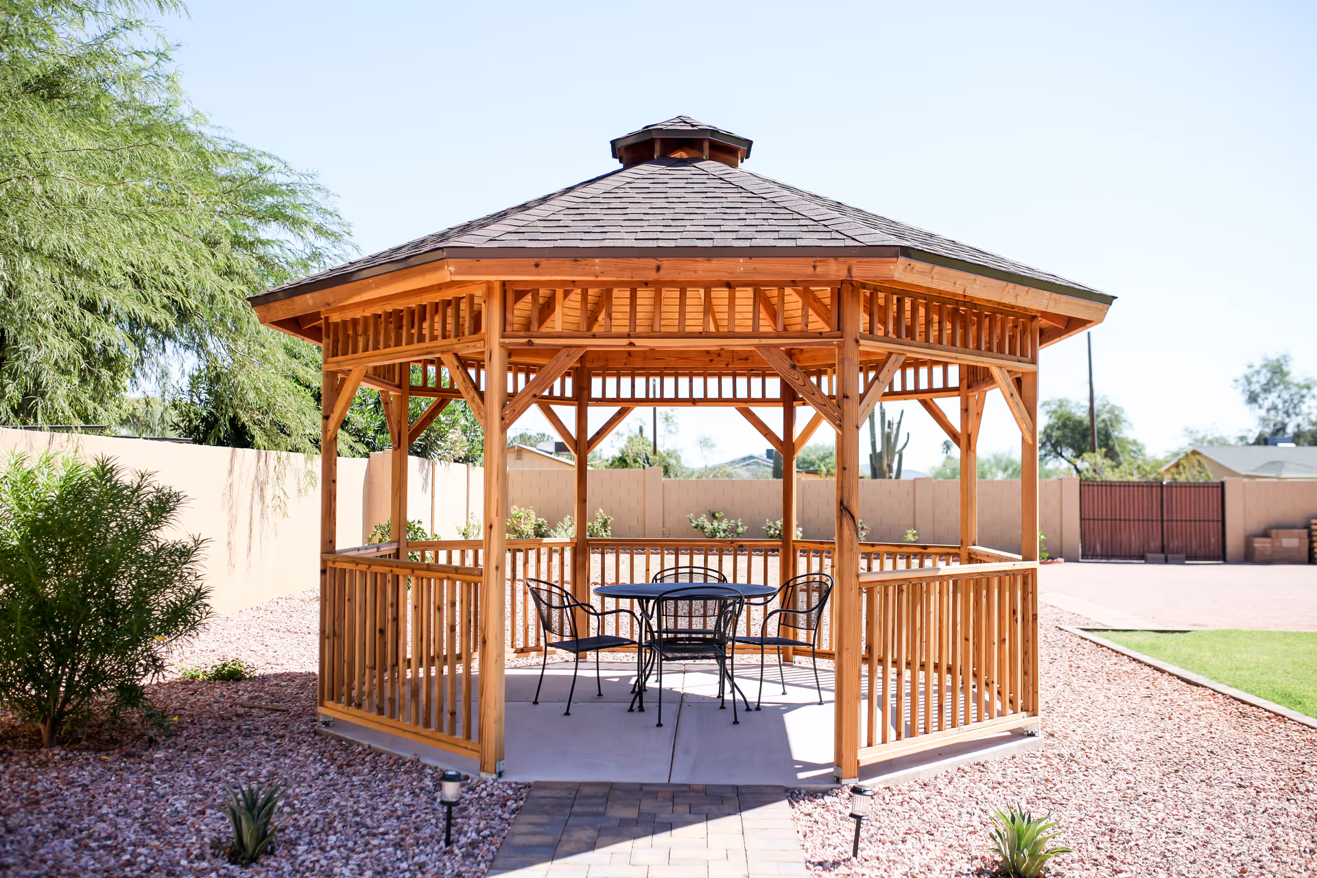 A wooden gazebo with a shingled roof in an outdoor garden area. Inside the gazebo, there is a round metal table with four matching chairs. The surrounding area has gravel, some small plants, and a beige wall with a gate in the background under a clear blue sky.