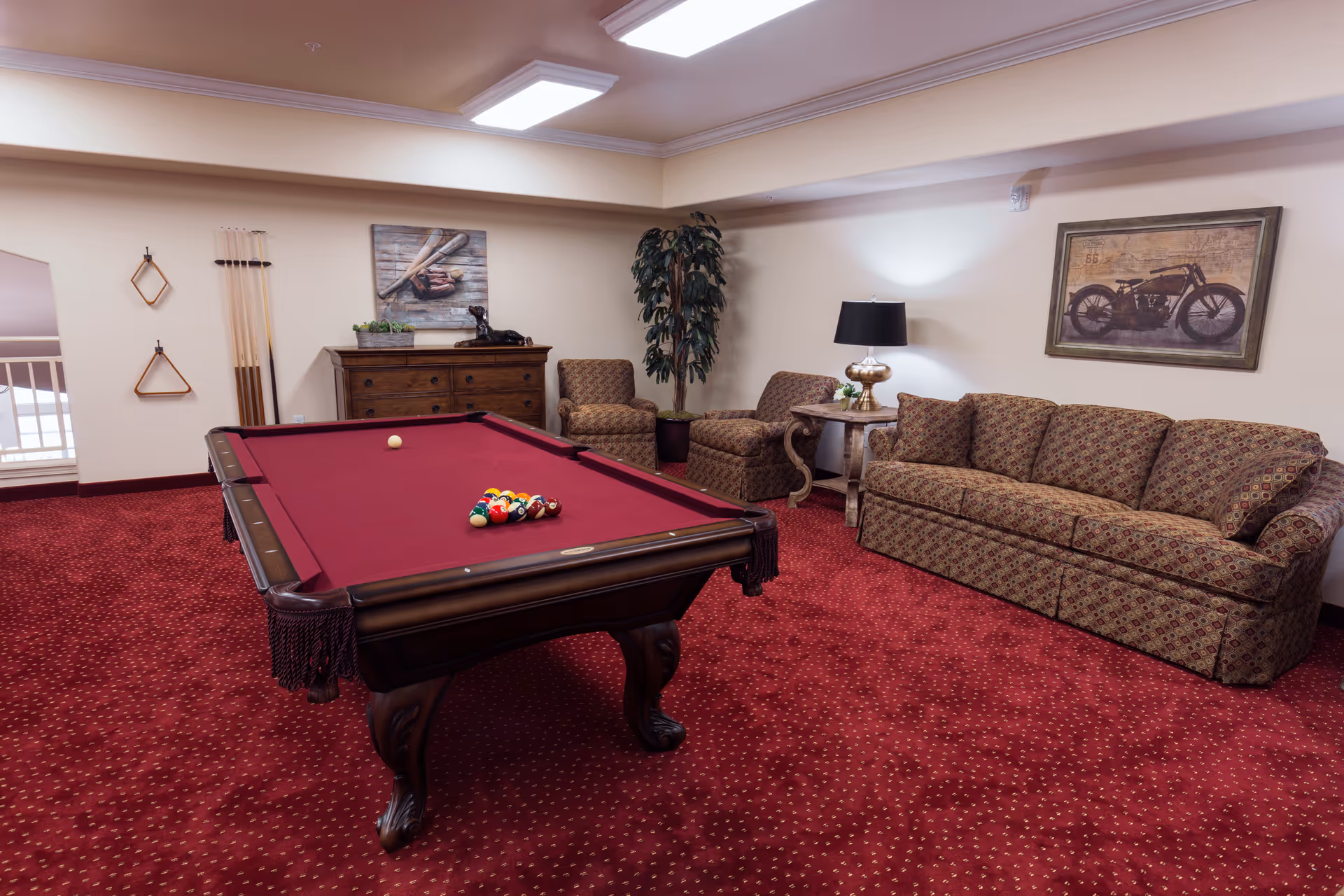 A cozy game room with a red-felt pool table in the center, a patterned sofa, two armchairs, a side table with a lamp, a wooden chest of drawers, and wall art including a motorcycle picture and a baseball bat and glove. The room has red carpet and beige walls.