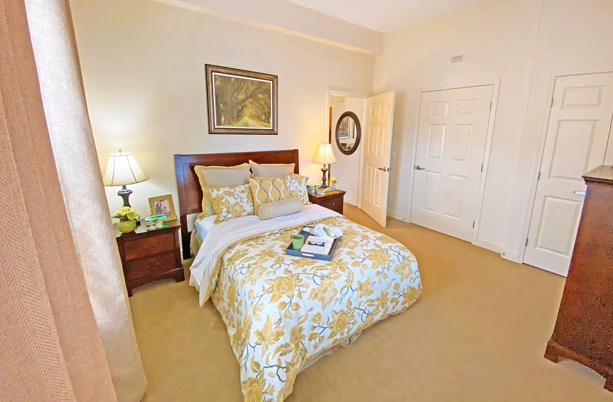 A bright bedroom with a bed covered in yellow and white floral bedding, two bedside tables with lamps, a framed picture on the wall above the bed, a round mirror on the wall near an open door, and beige carpeted floor.