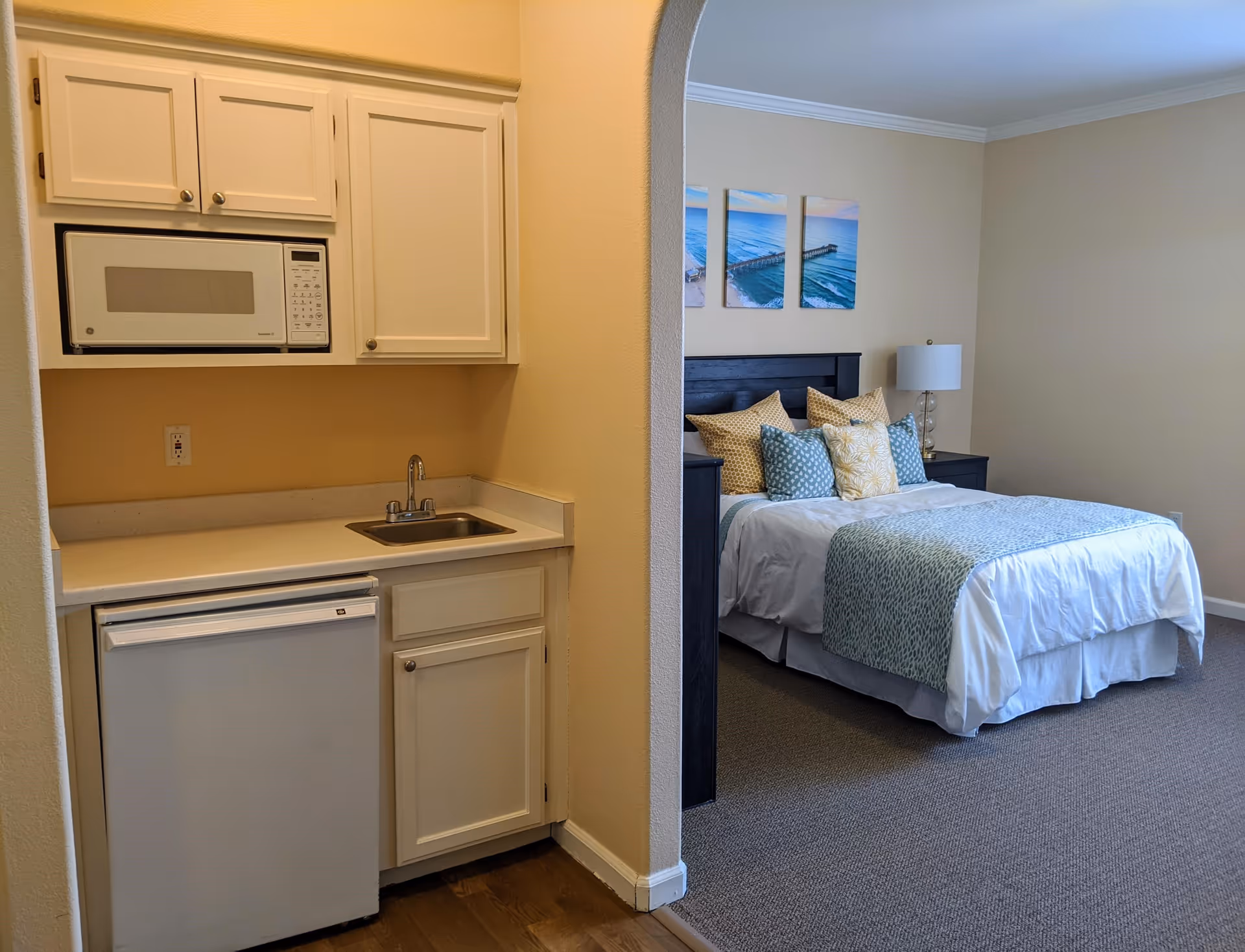 A small kitchenette with white cabinets, a microwave, a sink, and a mini refrigerator is visible on the left side. Through an arched doorway, a bedroom with a neatly made bed featuring multiple pillows and a blue patterned throw blanket is seen. Above the bed, there are three framed pictures of a pier over the ocean. A bedside table with a lamp is next to the bed.