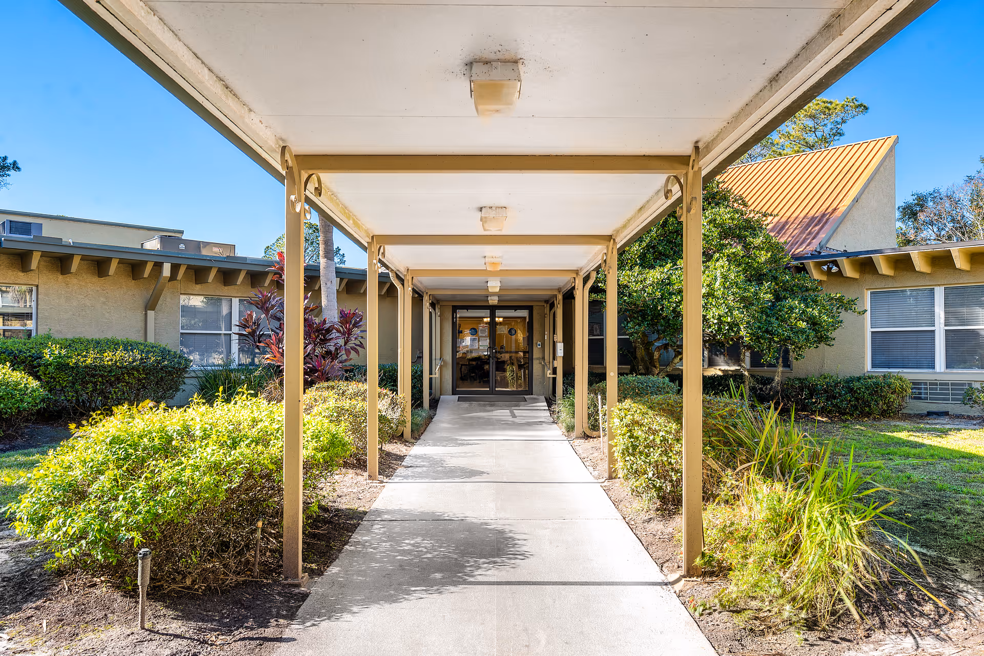 Covered walkway leading to the entrance of a single-story building with beige walls and a metal roof, surrounded by green bushes and trees under a clear blue sky.