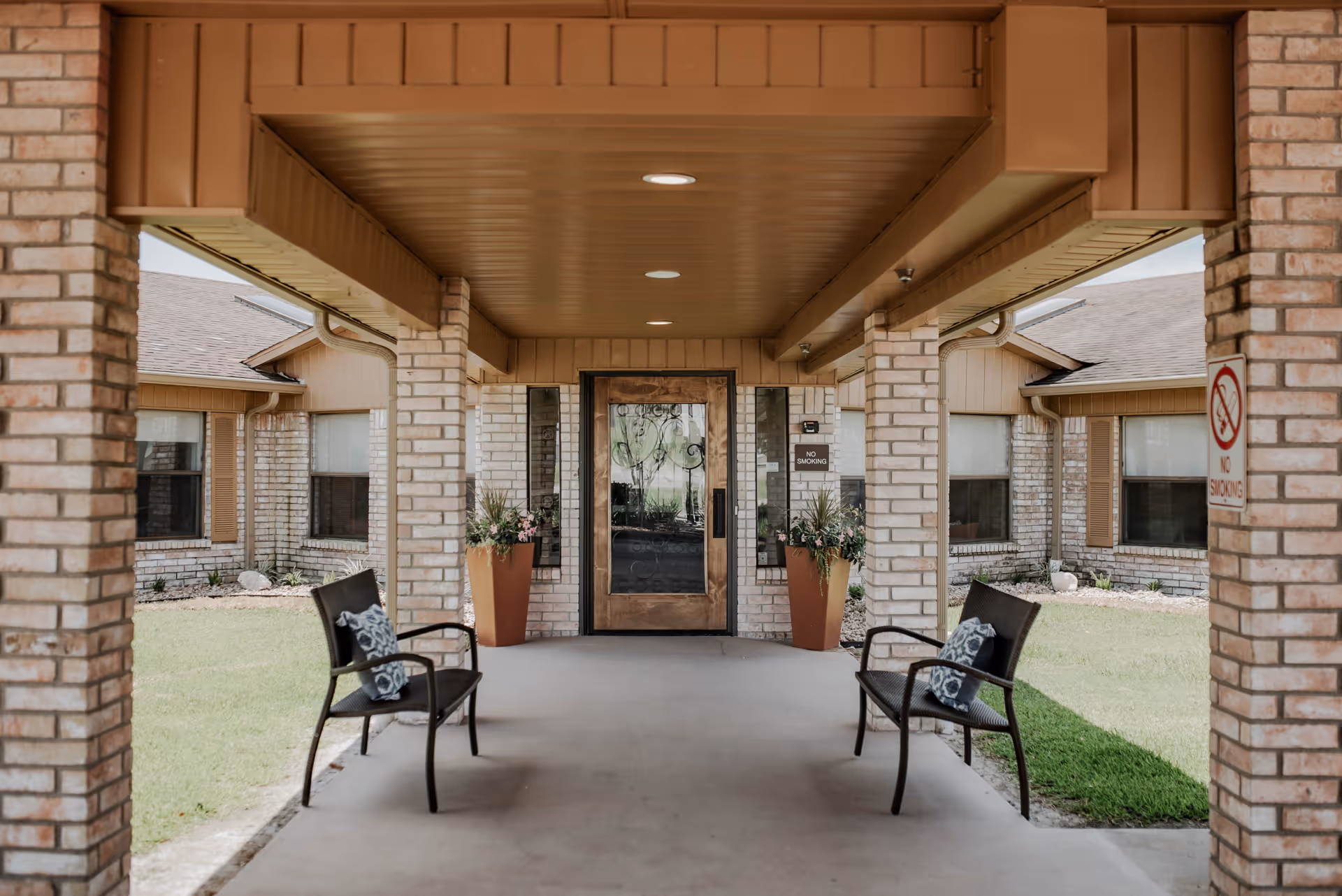 Covered entrance area of a building with brick pillars and a brown ceiling. Two black chairs with patterned cushions are placed on either side of the walkway. There are two tall planters with flowers near the glass door entrance. The building exterior is made of light-colored bricks with brown trim. A 'No Smoking' sign is visible on one of the pillars.