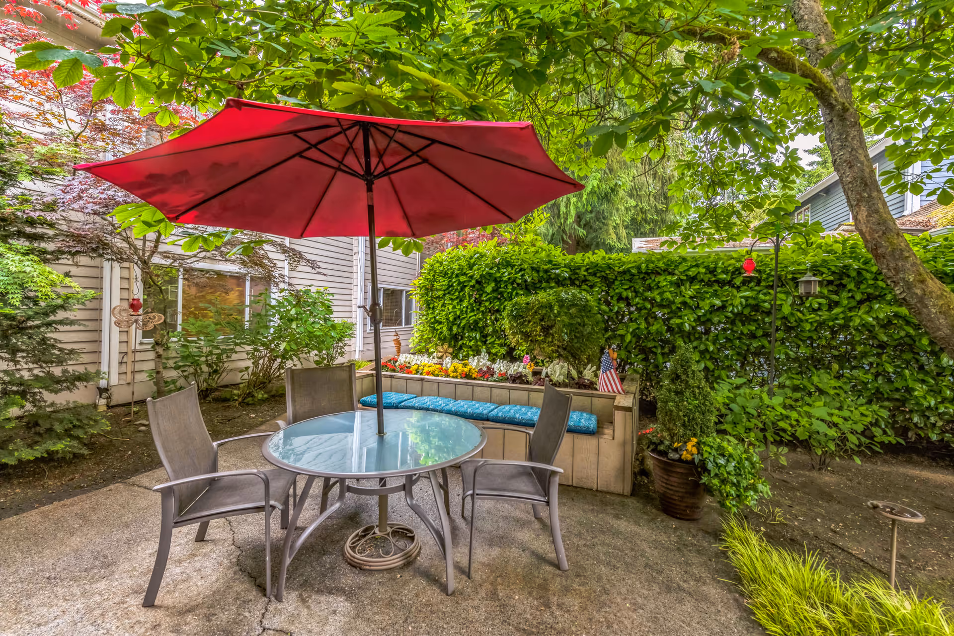 Outdoor patio area with a round glass table, four chairs, and a large red umbrella. The patio is surrounded by lush green plants, trees, and a hedge. There is a built-in bench with blue cushions and colorful flowers planted behind it. A small American flag is placed among the flowers.