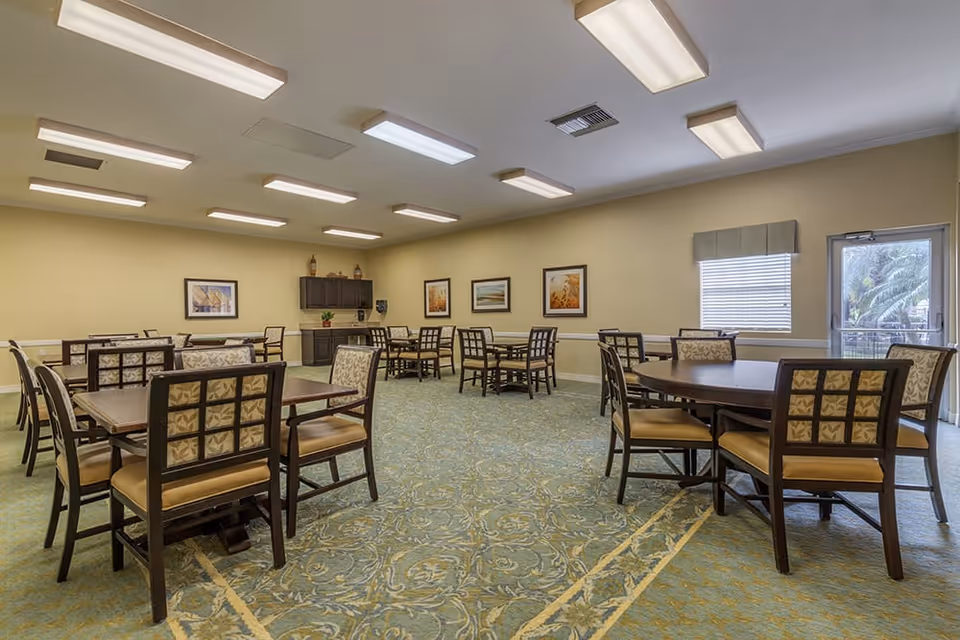 A spacious dining room with multiple wooden tables and cushioned chairs arranged neatly on a patterned carpet. The walls are painted beige and decorated with framed artwork. There is a window with blinds and a door leading outside. The ceiling has several rectangular fluorescent lights.