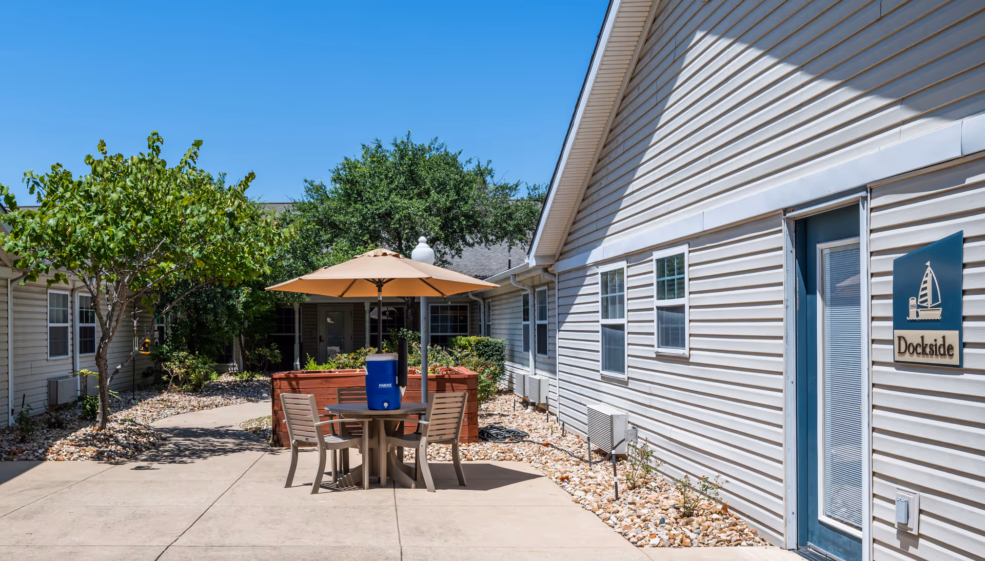 Outdoor courtyard area at Arden Courts - ProMedica Memory Care Community (Austin) featuring a round table with four chairs and a large umbrella. There is a blue water cooler on the table. The courtyard is surrounded by light-colored buildings with windows and a door labeled 'Dockside'. Trees and shrubs are planted along the edges with rocks covering the ground near the buildings. The sky is clear and blue.