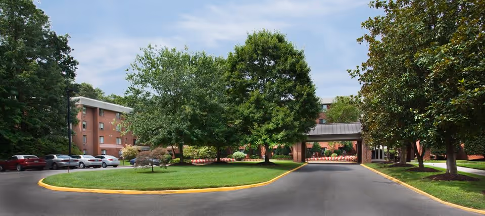 Entrance driveway to a senior living facility with a covered drop-off area, surrounded by green trees and well-maintained landscaping. Several parked cars are visible on the left side, and a multi-story brick building is in the background under a partly cloudy sky.