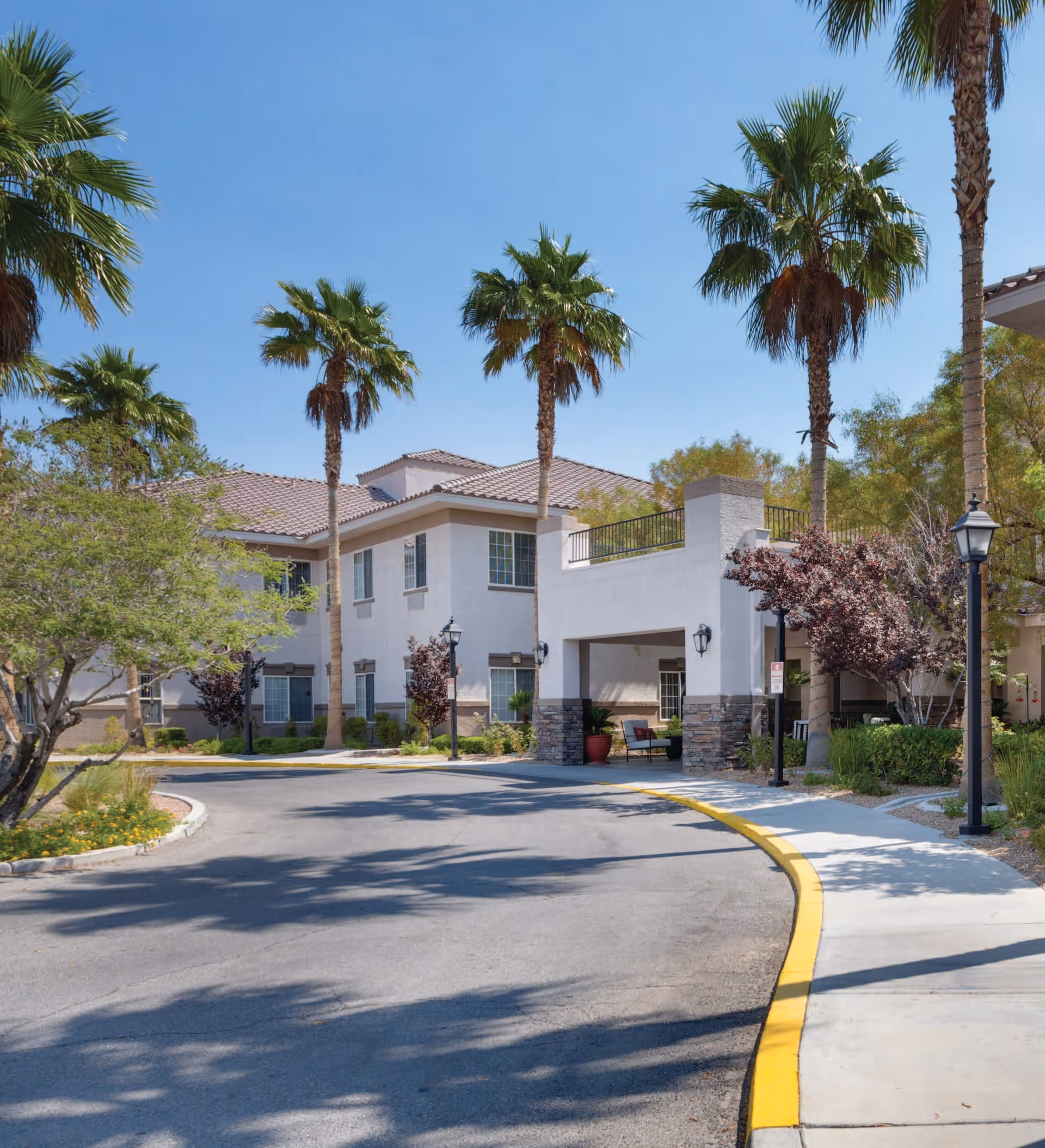 Exterior view of a senior living facility named Heritage Springs with a curved driveway, palm trees, and a two-story building with a covered entrance area. The sky is clear and blue.