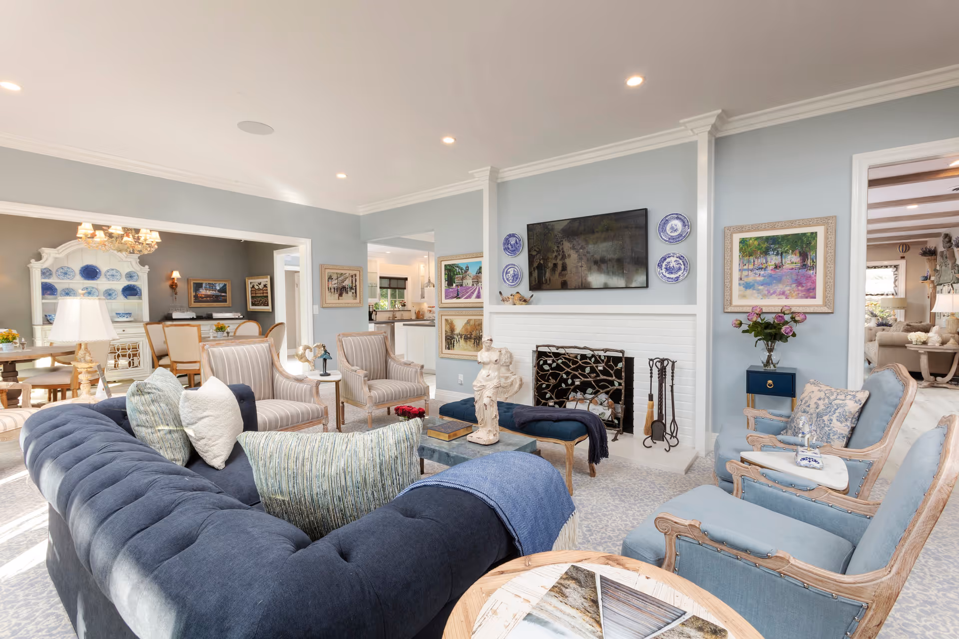 Bright, elegant living room with a navy tufted sofa, light blue armchairs, a fireplace with a mounted TV, and a dining area visible in the background.