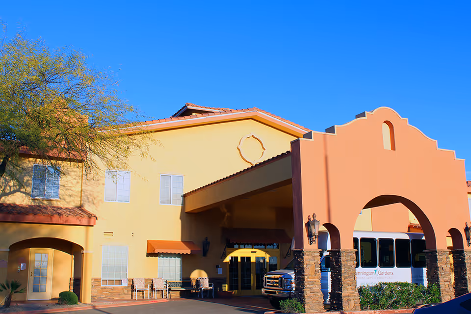 Exterior view of Pennington Gardens Assisted Living and Memory Care facility showing a yellow and orange building with a covered entrance and a white shuttle bus parked nearby under a clear blue sky.