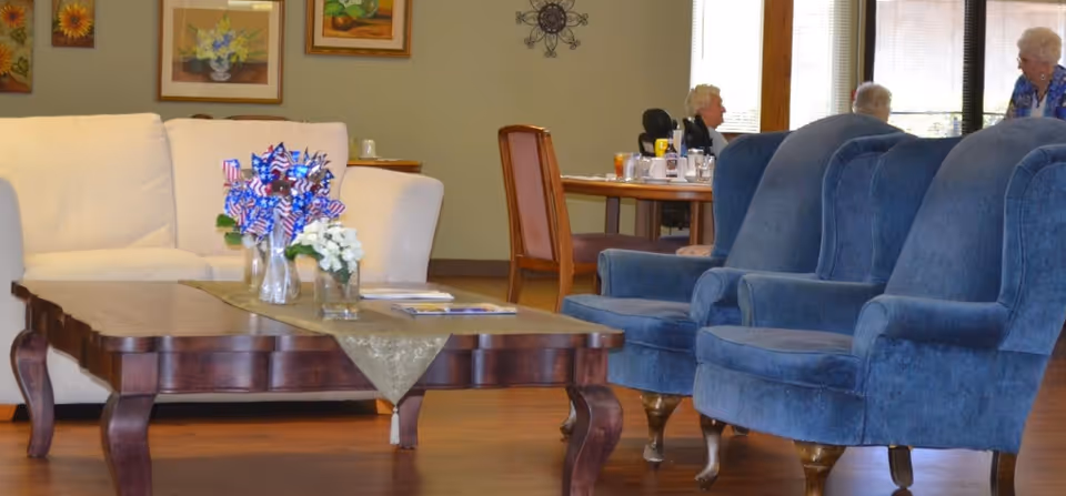 A senior living lounge with a wooden coffee table, cream sofa and blue armchairs, and residents seated at a table in the background.