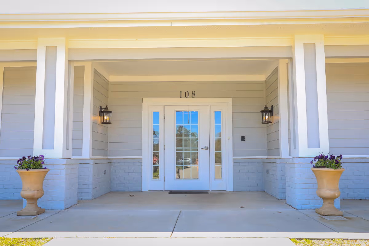 Entrance to a building with the number 108 above a white door with glass panels. The entrance is framed by two large columns and two wall-mounted lantern-style lights. There are two large planters with purple flowers on either side of the entrance.