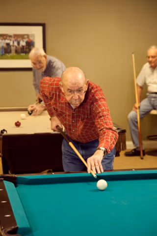 Three elderly men playing billiards in a recreational room. One man in a red plaid shirt is aiming to strike the cue ball with a pool cue, while another man in a gray shirt is preparing to take a shot on a second pool table in the background. A third man is sitting on a stool holding a pool cue, watching the game. A framed photo hangs on the beige wall behind them.