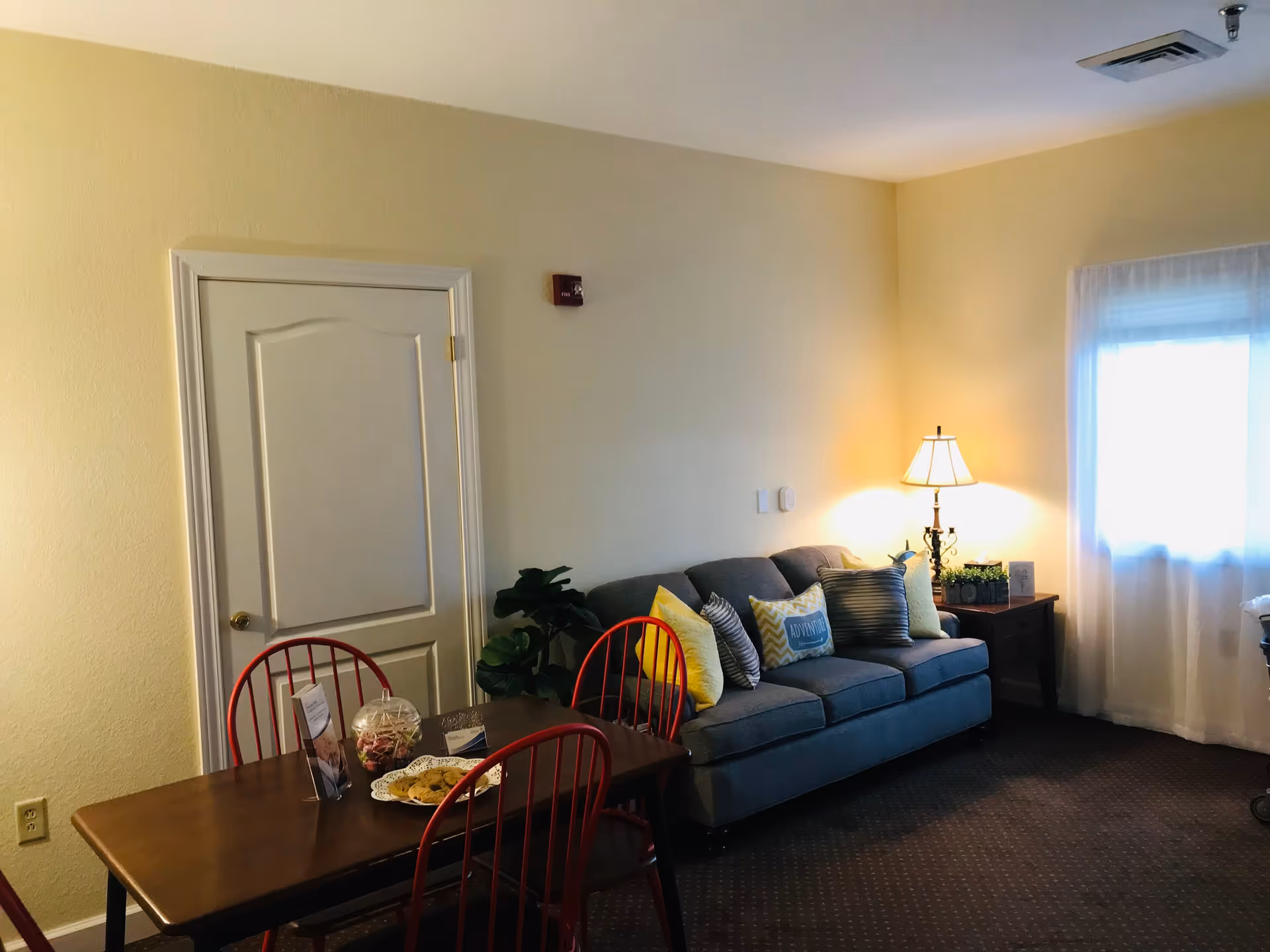 A cozy living area with a gray sofa adorned with multiple colorful pillows, a wooden side table with a lamp and decorative items, a window with sheer white curtains, and a wooden dining table with red chairs. The walls are painted light yellow and there is a closed white door on the left side.