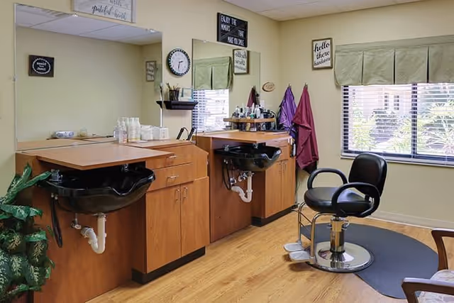 Interior view of a hair salon area with two black hair washing sinks set into wooden cabinets, a black salon chair on a circular mat, a window with green valance, and various hair care products and decor on the walls.
