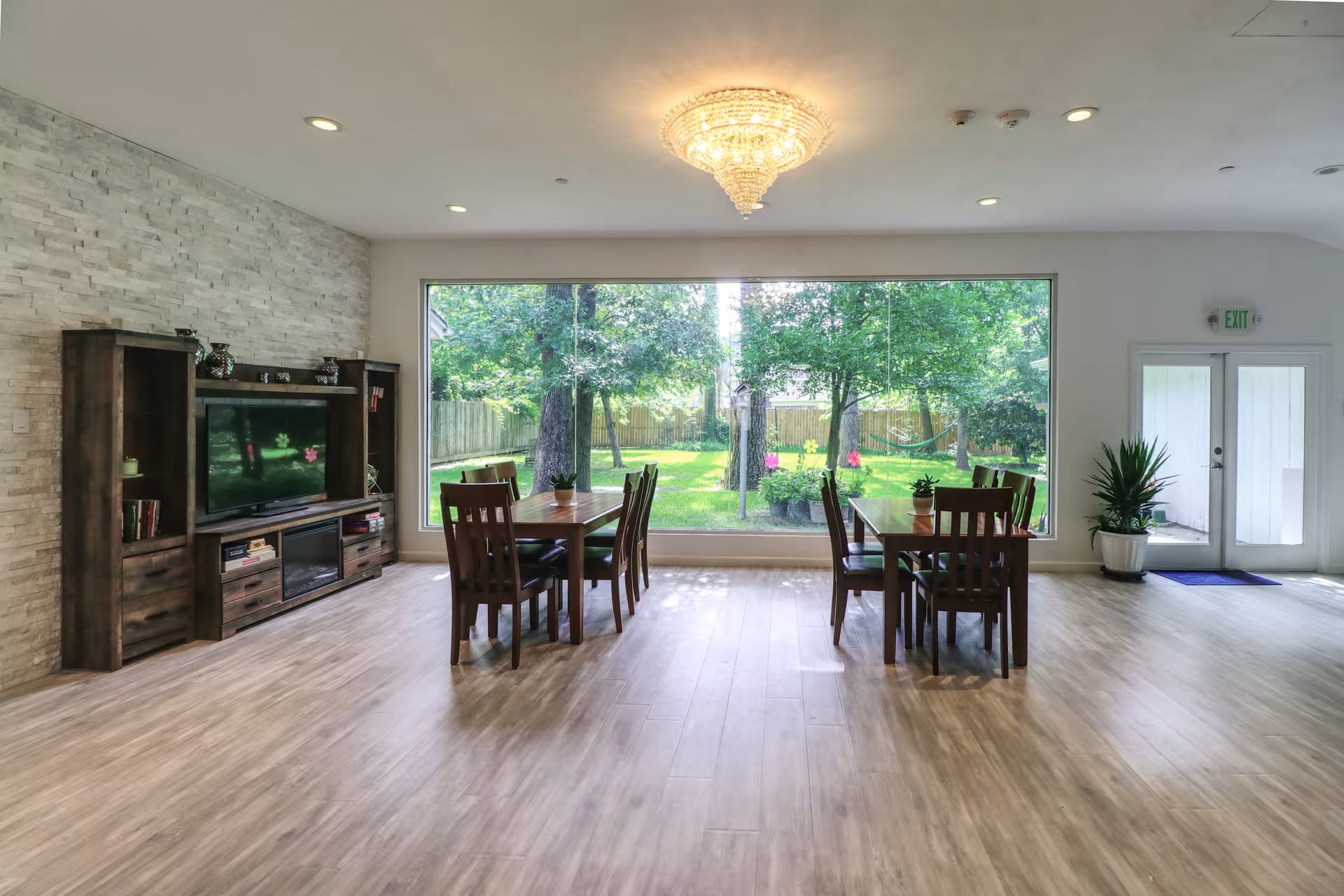 A bright and spacious common area with two wooden tables and chairs, a large window overlooking a green garden with trees, a wooden entertainment center with a TV, and a chandelier hanging from the ceiling. There is a door leading outside and potted plants near the entrance.
