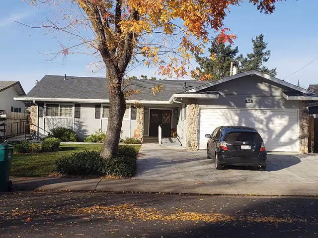 Single-story residential building with a two-car garage, driveway with a parked car, front lawn and a tree with autumn leaves.
