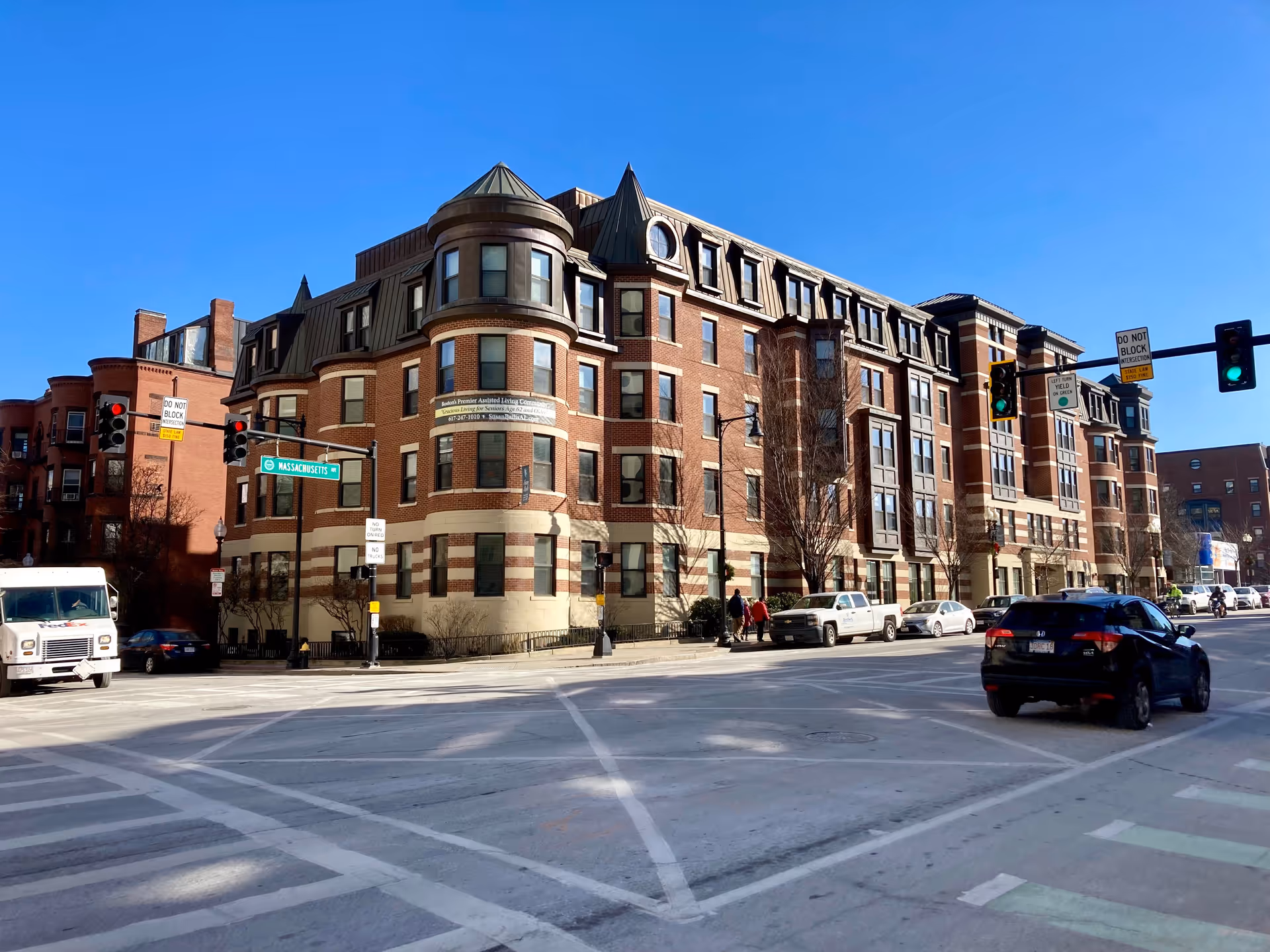 Brick multi-story assisted living building on a sunny street corner with traffic lights, cars, and pedestrians.