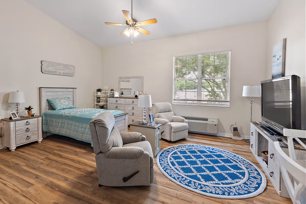 Bright assisted-living bedroom with a single bed, two recliners, dresser, TV, and a blue round rug.