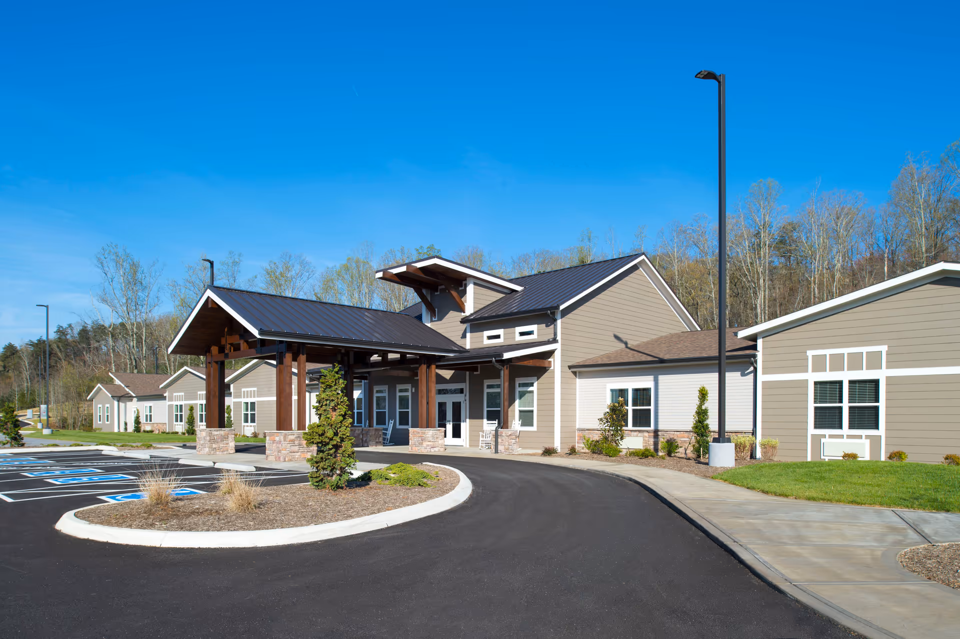 Exterior view of Pinnacle Assisted Living facility showing a modern building with a covered entrance, multiple windows, a paved driveway, parking spaces including handicapped spots, and surrounding greenery under a clear blue sky.