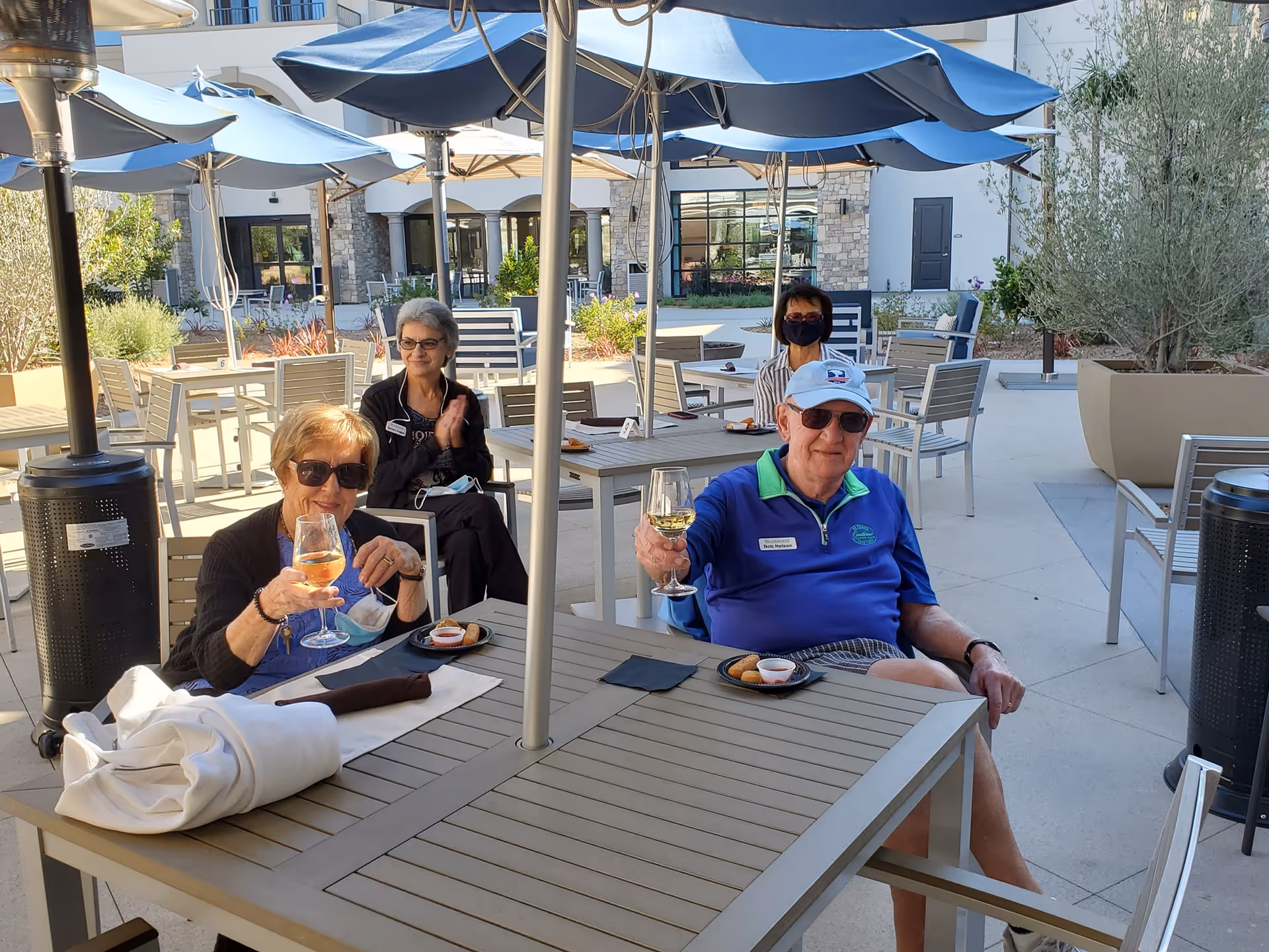 Three elderly people sitting at outdoor tables under blue umbrellas at Silvergate Rancho Bernardo. Two of them are holding glasses of white wine and smiling, while the third person is sitting behind them clapping. The area is a patio with modern outdoor furniture and potted plants.