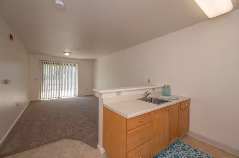 Interior view of a senior living apartment showing a small kitchen area with wooden cabinets, a sink, and a countertop. Adjacent to the kitchen is a carpeted living room area with a large sliding glass door covered by vertical blinds, allowing natural light to enter the space.