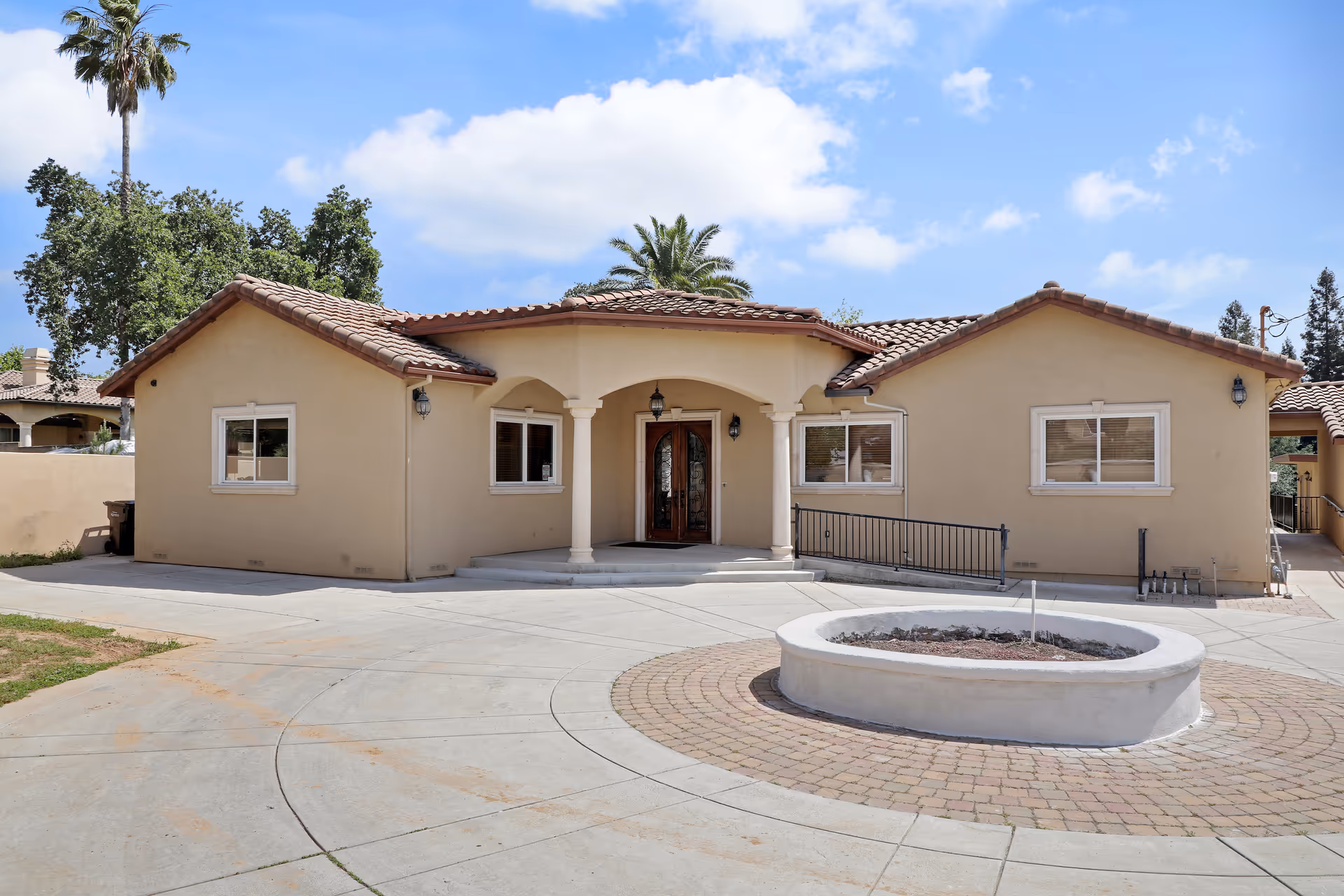 Exterior view of a single-story beige stucco building with a tiled roof, featuring a central entrance with double wooden doors under an arched porch supported by two columns. There is a circular paved area with a raised planter in front of the building, and palm trees and other greenery in the background under a partly cloudy sky.