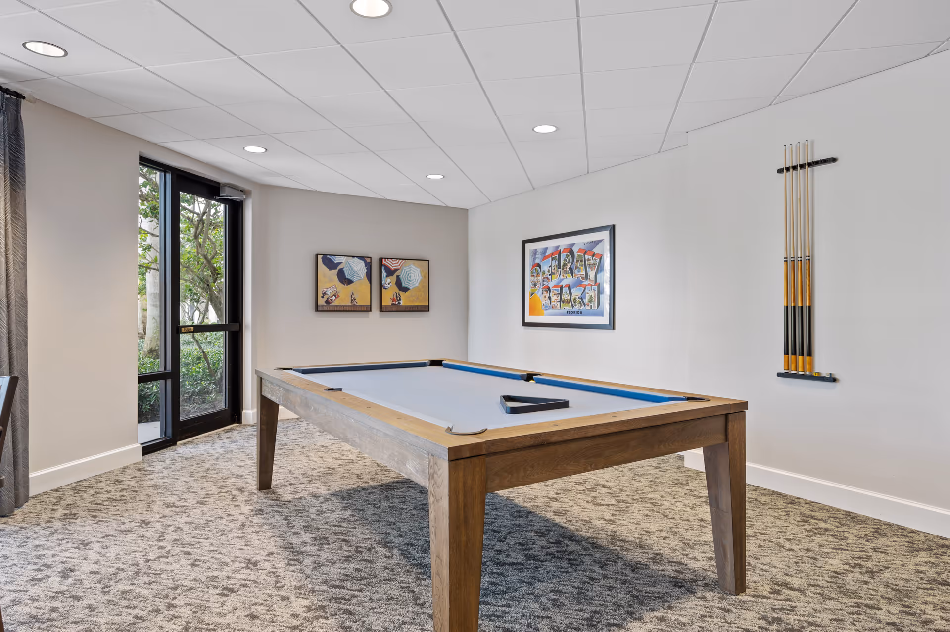 A bright room with a pool table featuring a light blue felt surface and wooden frame. On the wall to the right, there is a rack holding five pool cues. The walls are decorated with three framed artworks, including one that says 'Delray Beach Florida'. A glass door with a black frame leads outside, and the floor is covered with patterned carpet.