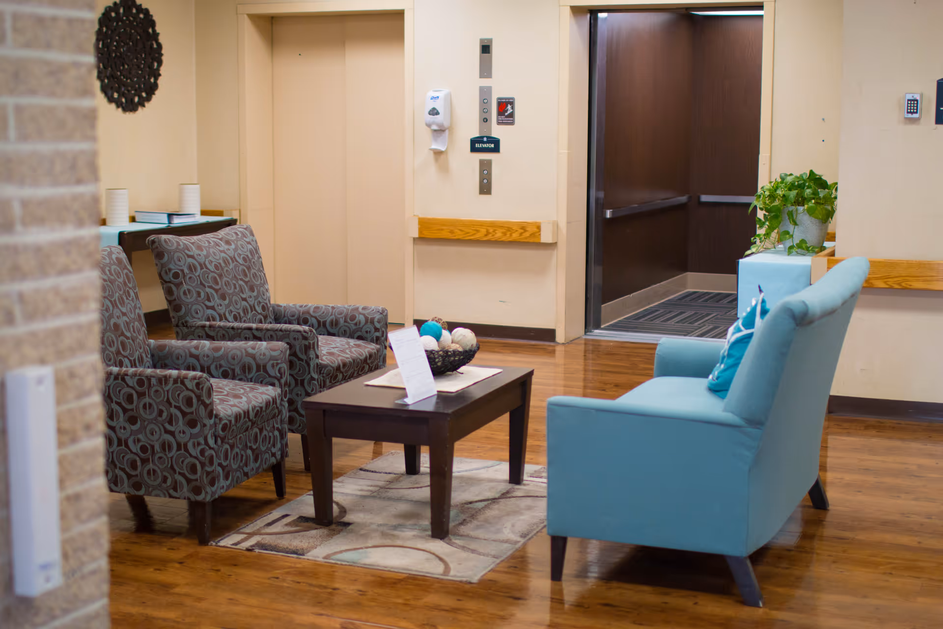 A seating area in a senior living facility with two patterned armchairs and a light blue loveseat arranged around a dark wooden coffee table on a rug. Behind the seating area, there is an elevator with beige doors and a small table with books and cups. A potted plant is placed on a small table near the elevator.