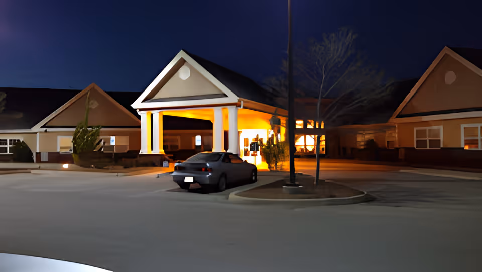 Night view of the entrance to a senior living facility with a covered drop-off area illuminated by warm lights. A car is parked near the entrance, and the building has a peaked roof design with beige and brown exterior walls. There are some small trees and a streetlamp in the parking area.