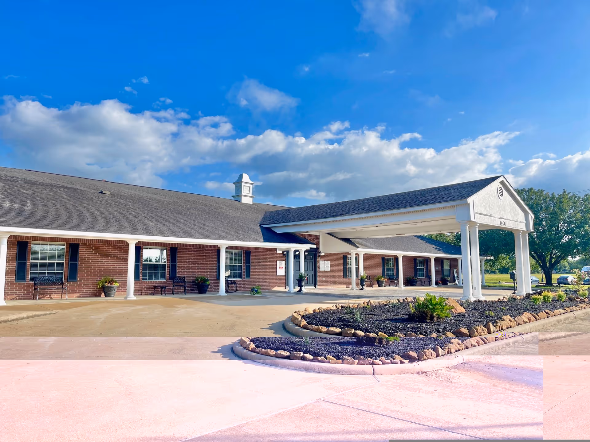 Front exterior of a single-story brick senior living facility with a covered porte-cochere and landscaped driveway under a blue sky.