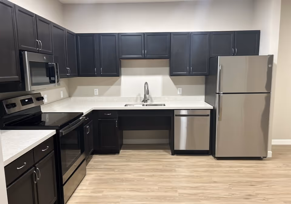 Modern kitchen with dark cabinetry, stainless steel appliances including a refrigerator, dishwasher, microwave, and electric stove, white countertops, and light wood flooring.