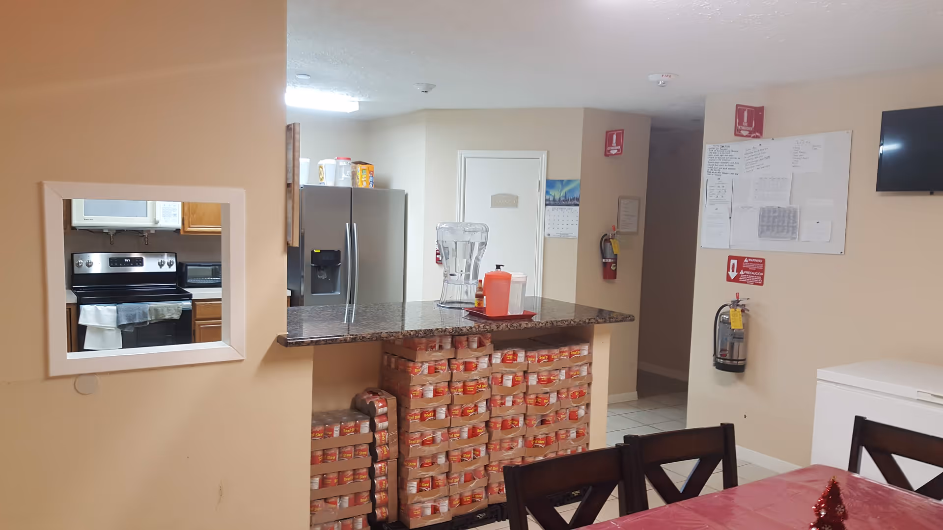 Communal dining area with a countertop and stacked canned goods beneath it, refrigerator and stove visible through a pass-through, and chairs around a table.