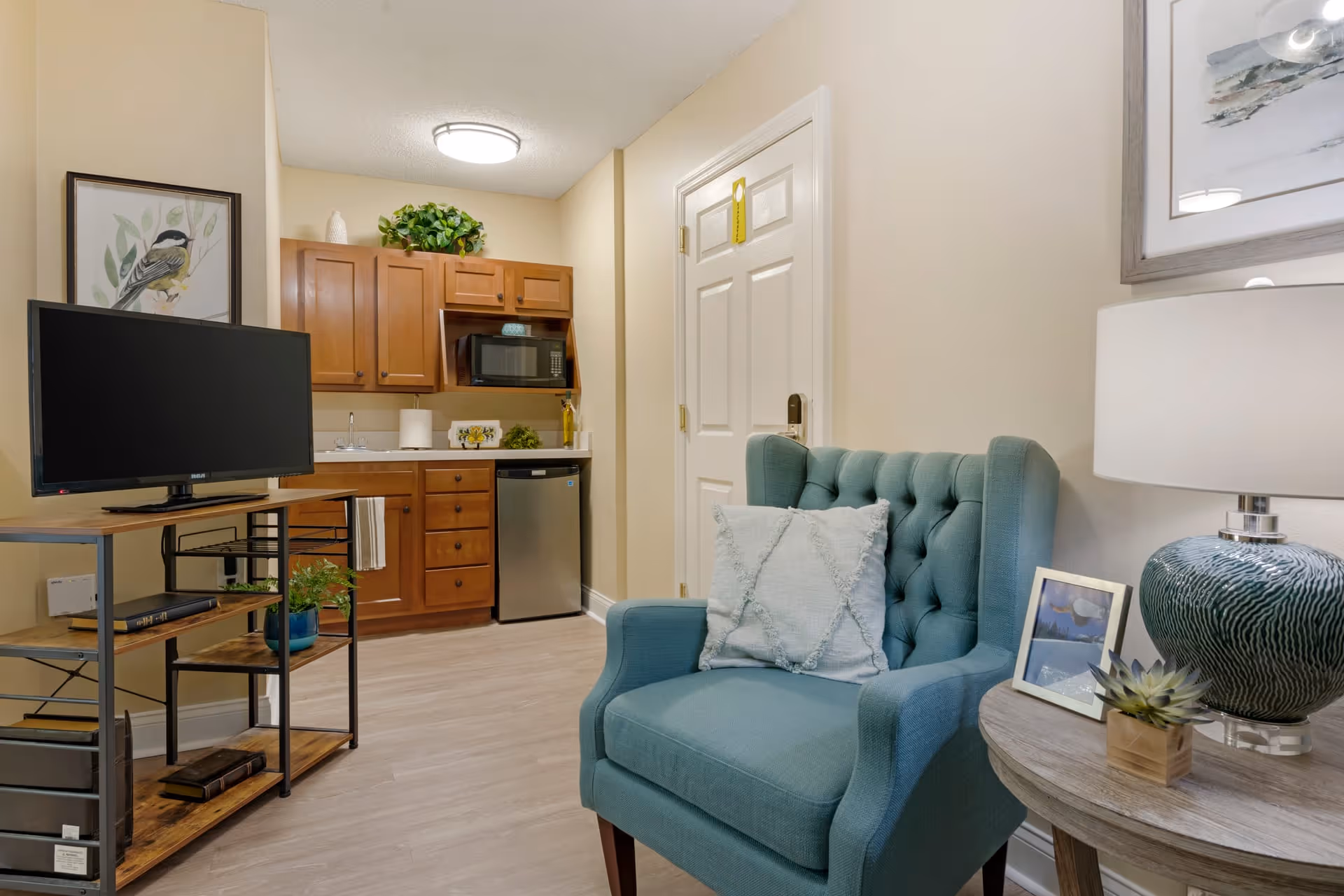 A cozy interior space featuring a teal upholstered armchair with a decorative pillow, a round wooden side table with a lamp, a small plant, and a framed photo. In the background, there is a kitchenette with wooden cabinets, a microwave, a small refrigerator, and a TV on a metal and wood stand. The walls are light beige, and there is a framed bird artwork on the wall.