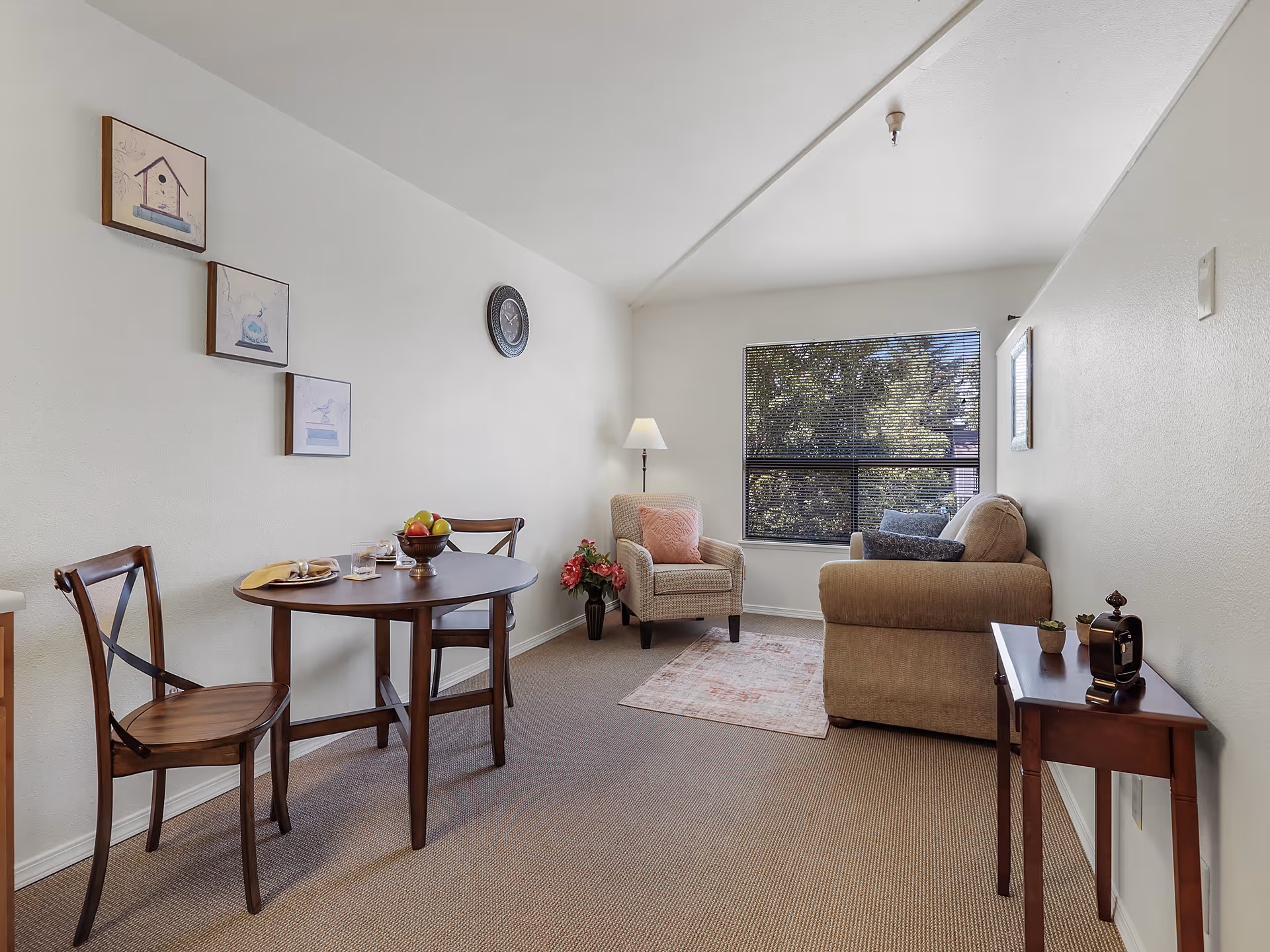 A cozy living area in an assisted living facility featuring a small round wooden dining table with two chairs, a beige armchair with a pink cushion, a beige sofa with blue cushions, a floor lamp, a small wooden side table with decorative items, and a window with blinds showing trees outside. The walls are decorated with three framed pictures and a clock.
