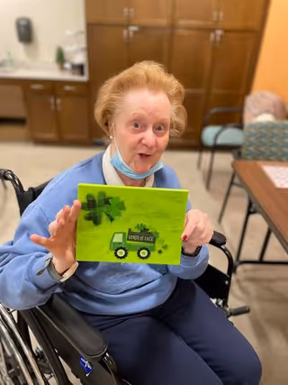 An elderly woman sitting in a wheelchair inside a room, holding a green craft board with a truck and shamrocks that says 'Loads of Luck'. She is wearing a blue sweatshirt and has a face mask pulled down below her chin. Behind her are wooden cabinets and chairs around a table.