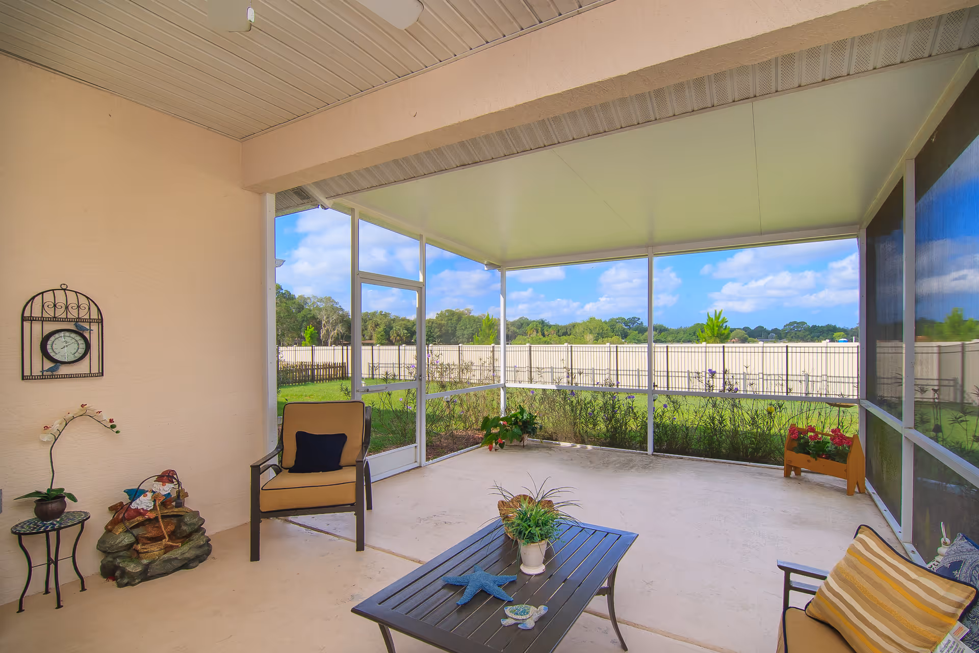 A screened-in patio area with beige walls and ceiling, featuring two cushioned chairs with pillows, a black coffee table with decorative items including a small plant and a starfish, a small side table with a potted plant, a decorative wall clock, and a small water fountain. Outside the screened area, there is a green lawn and a white fence under a blue sky with scattered clouds.