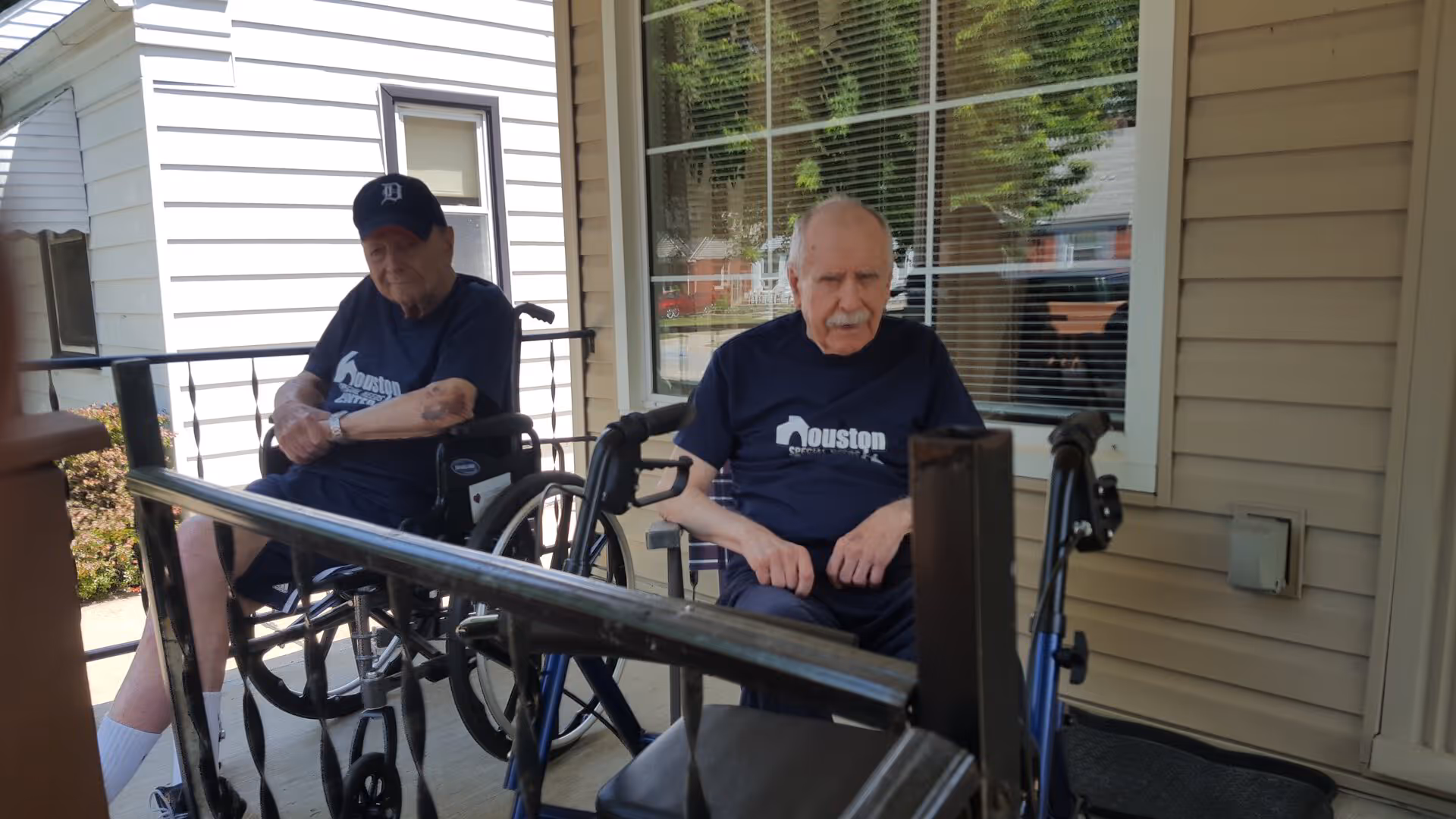 Two elderly men in wheelchairs sitting on a covered porch outside a beige-sided senior living building.