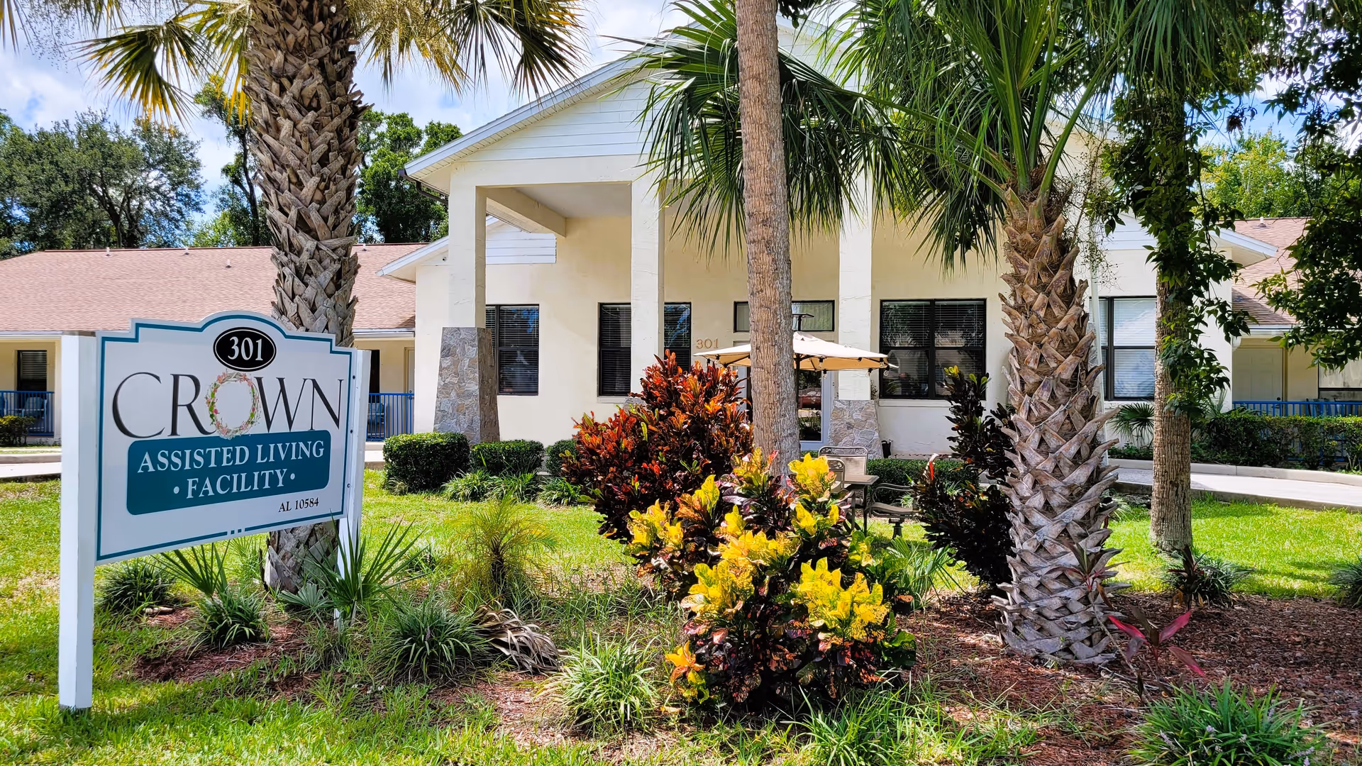 Exterior view of Crown Assisted Living Facility showing a white building with a covered entrance, surrounded by palm trees and colorful bushes. A sign in front displays the facility name and address number 301.