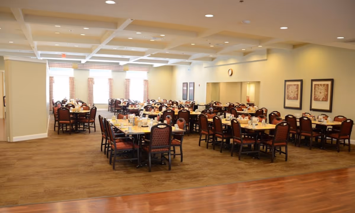 A spacious dining room in a senior living facility with multiple rectangular tables set with cups, plates, and utensils. The room has wooden flooring, beige walls, and several windows with checkered curtains allowing natural light to enter. There are framed pictures on the walls and a clock above a serving window.