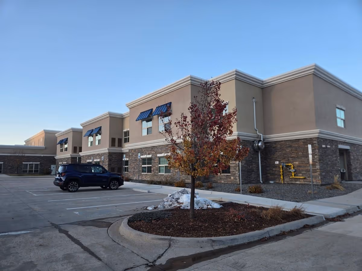 Exterior front of a two-story beige senior living building with stone accents, a parked SUV, and a small tree with autumn leaves in the parking lot.