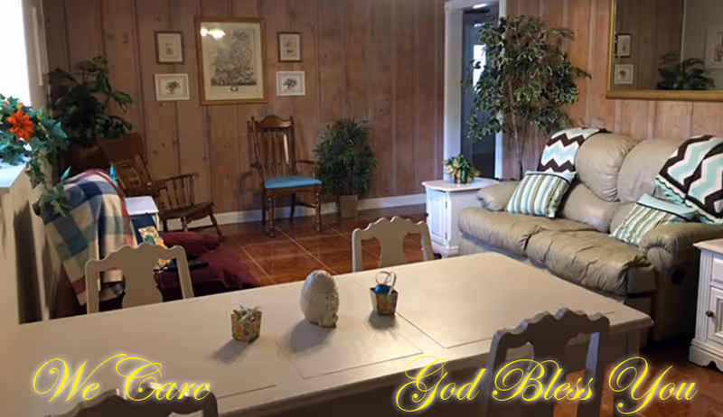 A cozy living room with wood-paneled walls, a beige leather sofa with patterned pillows, a white dining table with chairs, and various plants and framed pictures decorating the space.
