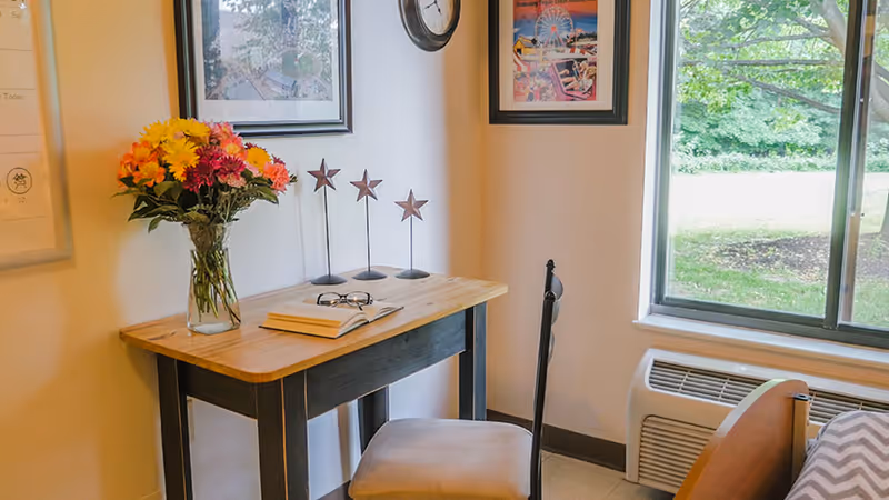 A small wooden desk with a vase of colorful flowers, an open book, and a pair of glasses on it. Behind the desk are three decorative star-shaped ornaments. A chair is placed in front of the desk. On the wall above the desk are two framed pictures and a round clock. To the right, there is a window showing green trees outside and an air conditioning unit below the window.