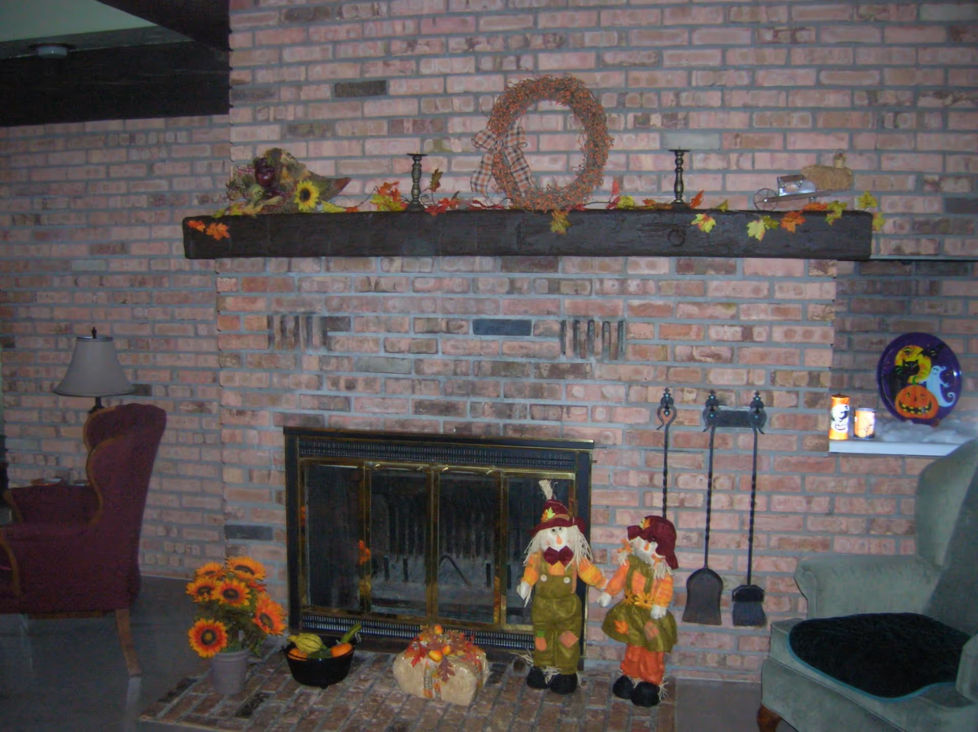 A cozy living room area featuring a brick fireplace decorated with autumn-themed items including a wreath, candles, and small scarecrow dolls. There are two armchairs, one burgundy and one green, positioned on either side of the fireplace. A small table lamp is visible next to the burgundy chair.