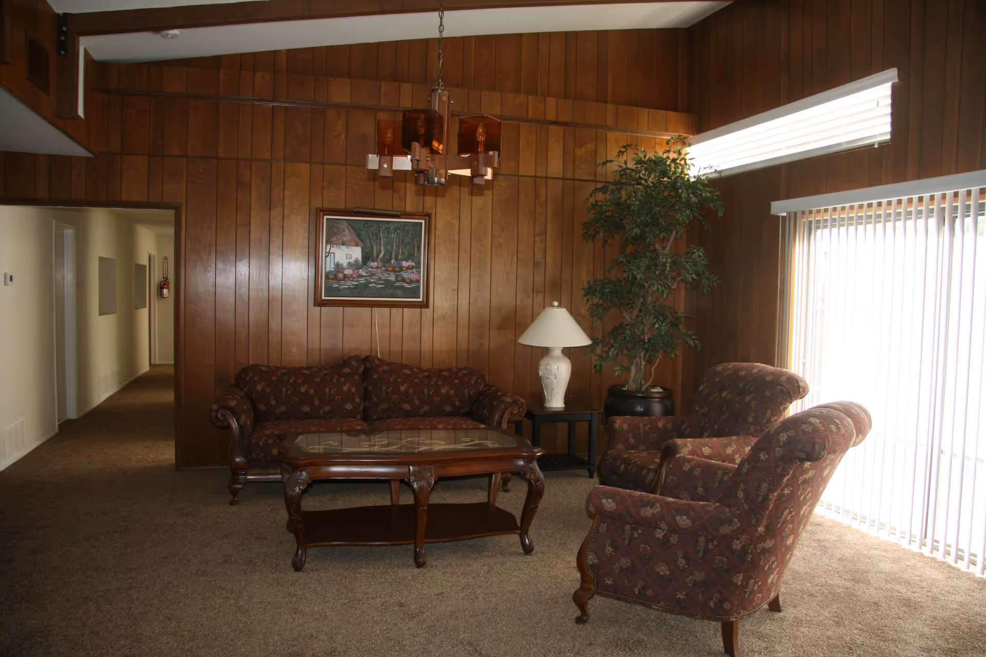 Wood-paneled living room with a sofa, two upholstered armchairs, coffee table, lamp and a potted plant next to vertical blinds.