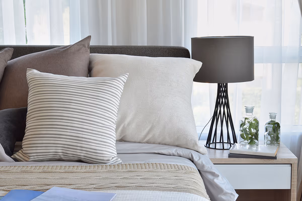 Close-up of a neatly made bed with multiple pillows including a striped pillow, a beige pillow, and a brown pillow. Next to the bed is a wooden nightstand with a modern black lamp, two glass bottles with green leaves inside, and a closed book. Sheer white curtains cover the window in the background.