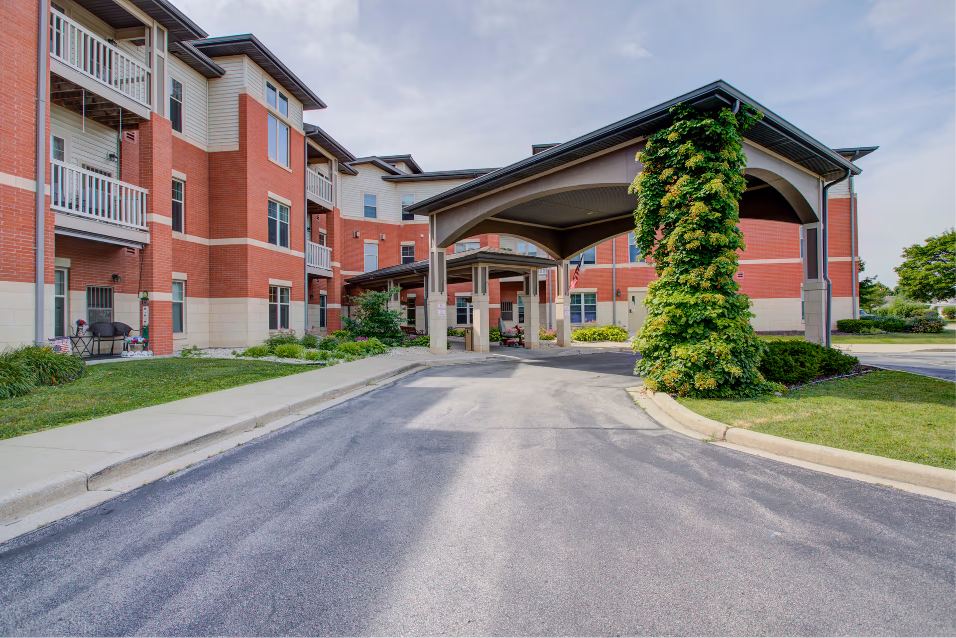 Exterior view of Dimensions Living Green Bay facility showing a red brick building with white trim and balconies. There is a covered driveway entrance with a large archway partially covered in green ivy. The surrounding area includes a paved road, sidewalks, and landscaped greenery under a partly cloudy sky.