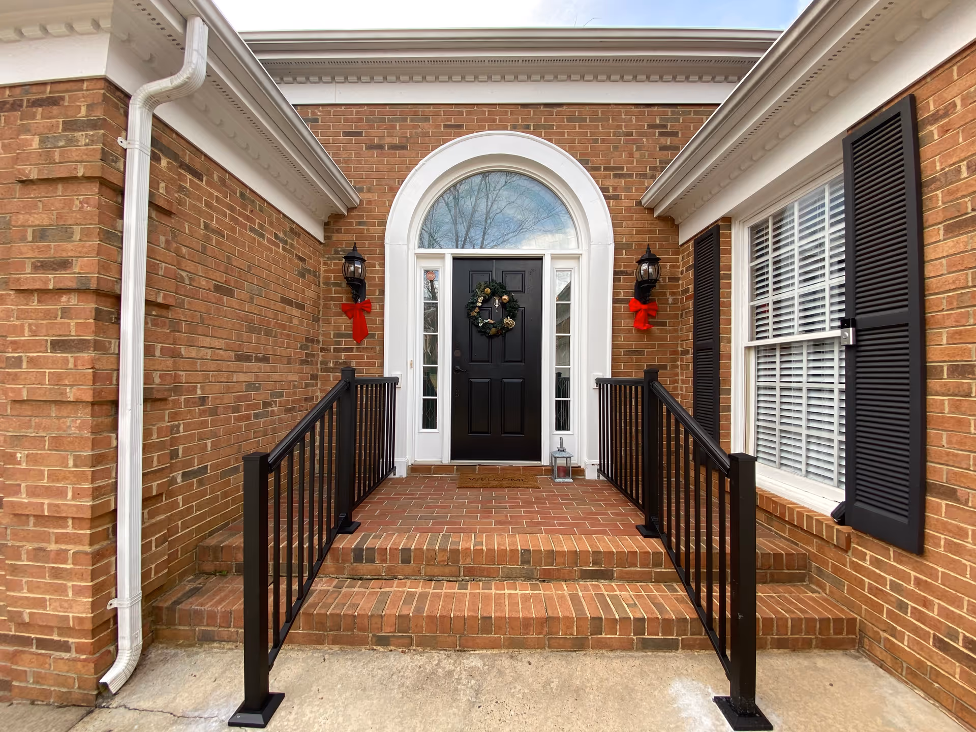 Brick building front entrance with a black door decorated with a wreath, flanked by lanterns with red bows and brick steps with black railings.