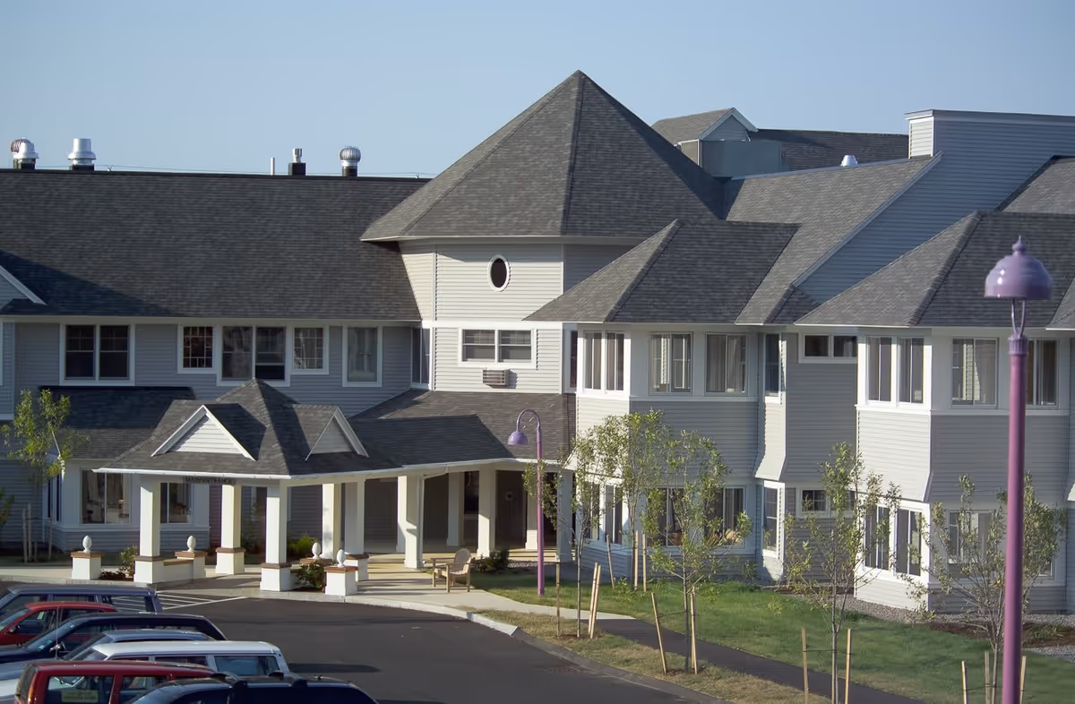 Exterior view of a large, multi-story retirement village building with gray shingled roofs and white siding. There is a covered entrance with columns and a driveway leading to it. Several small trees and a purple street lamp are visible in the landscaped area near the building. Several parked cars are visible in the foreground.
