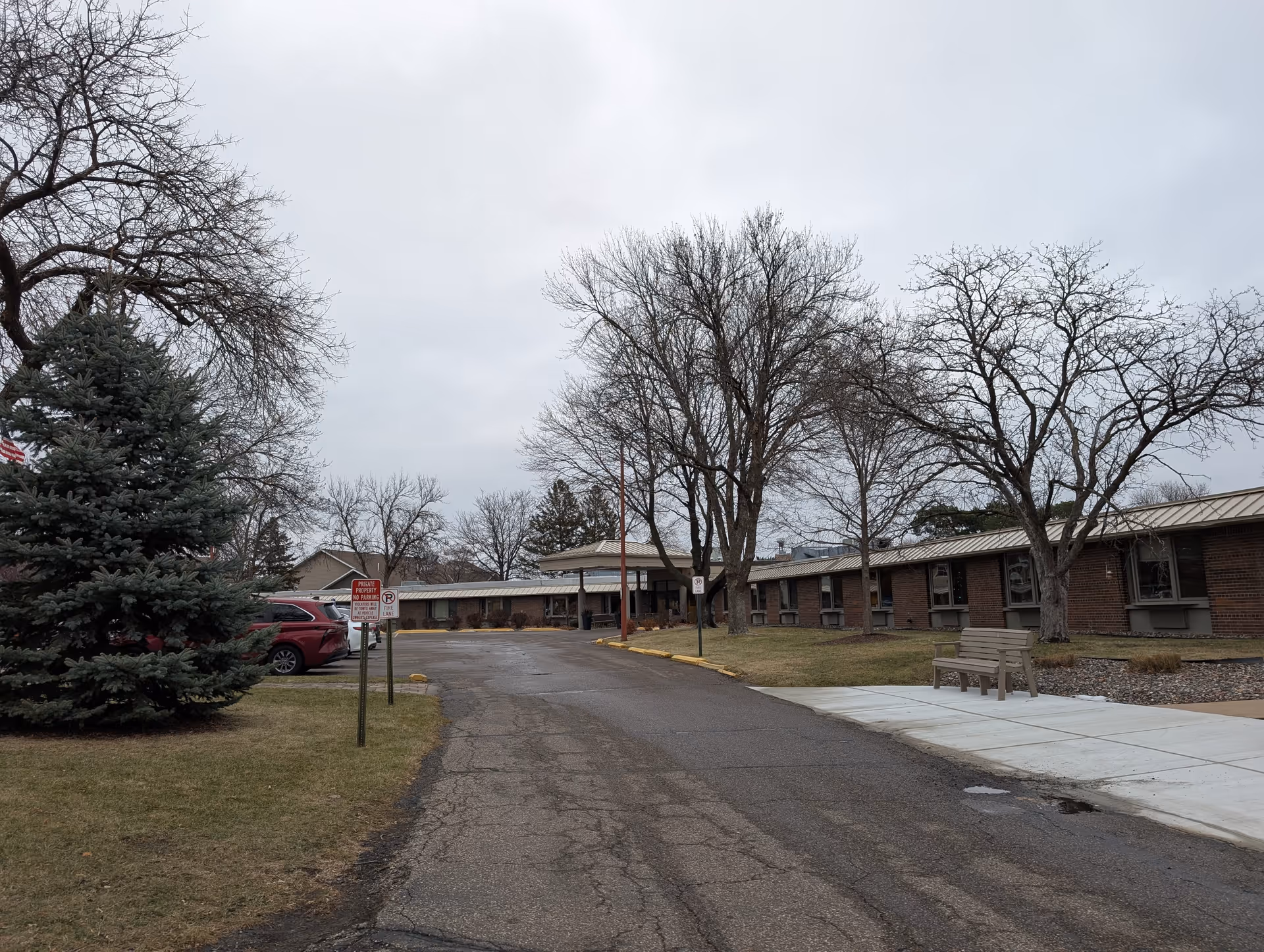 Exterior view of Good Samaritan Ambassador facility showing a single-story brick building with a metal roof, leafless trees, a bench on a sidewalk, a driveway, and parked cars under a cloudy sky.