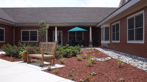 Outdoor courtyard area of a senior living facility with a wooden bench, landscaped garden beds with mulch and small plants, and a patio table with a teal umbrella. The building surrounding the courtyard has red siding and multiple windows.